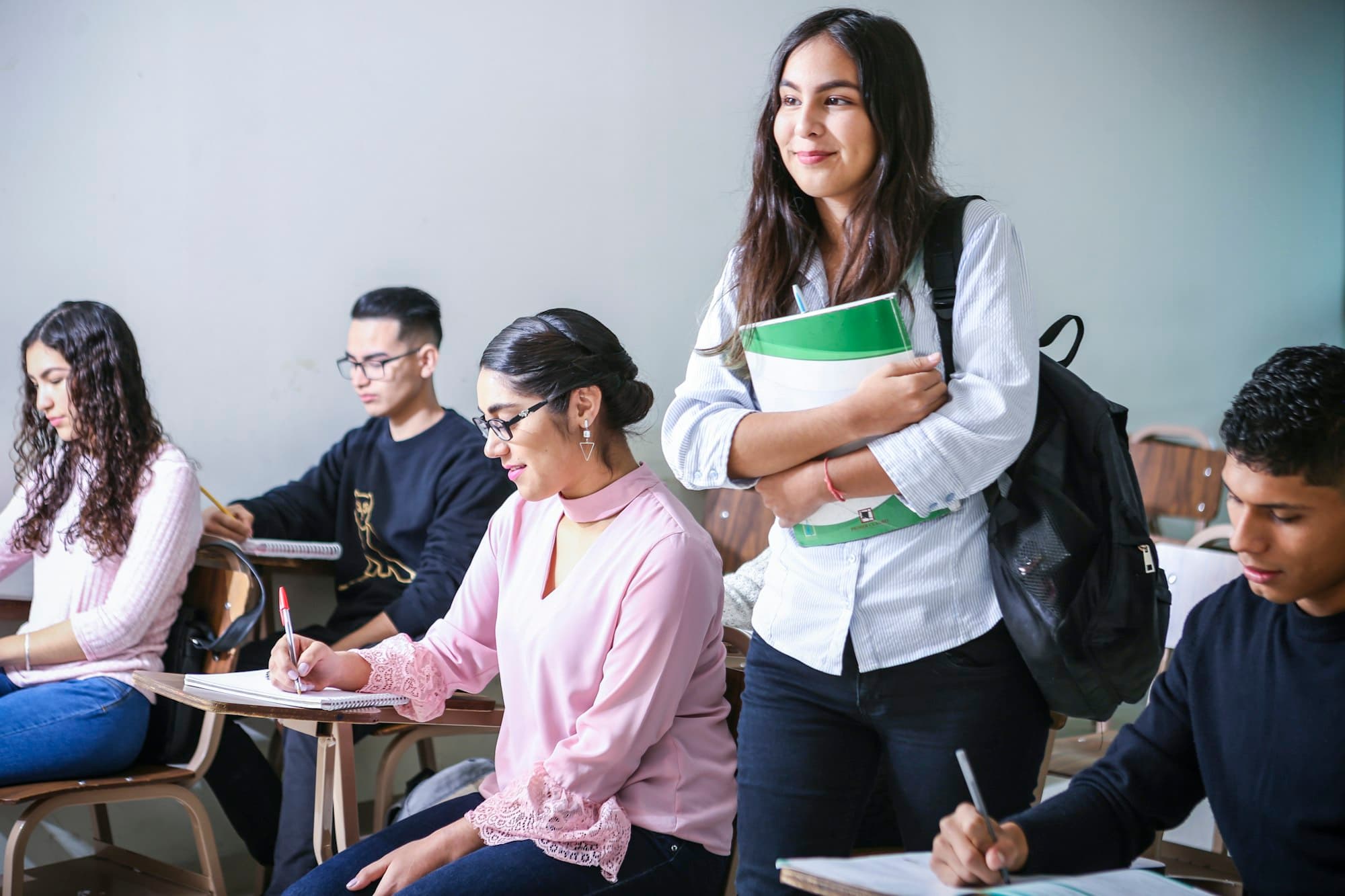 Students in a classroom, one standing and smiling while holding a stack of folders